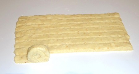 Охлаждённое тесто раскатать в пласт толщиной 5 мм, длиной примерно 30 см и нарезать на полоски шириной 1,5 см. Каждую полоску свернуть в рулет не до конца.
