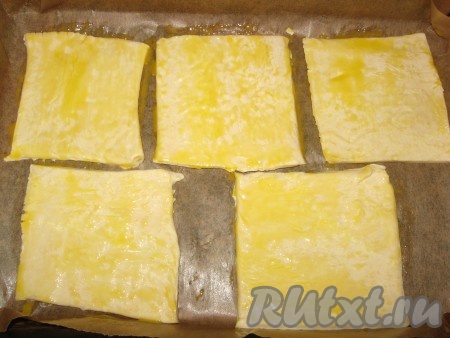 Заранее разогреть духовку до 220 градусов. Размороженное тесто разрезать на одинаковые части - квадраты или прямоугольники, выложить их на противень, застеленный пергаментом, смазать желтком.
