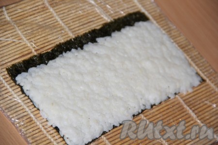 Разрезать лист нори на две половинки. На циновку выложить половину листа нори блестящей стороной вниз. Равномерно выложить рис на лист нори, оставляя один край нори на 1 см свободным от риса (как на фото). Толщина слоя риса должна быть не более 5 мм.
