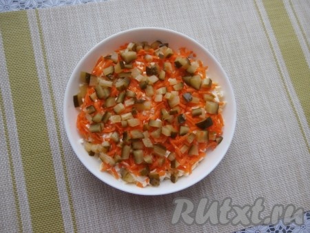 Затем выложить слой натёртой моркови. Поверх морковки разложить маринованный огурец, нарезанный на маленькие кубики.