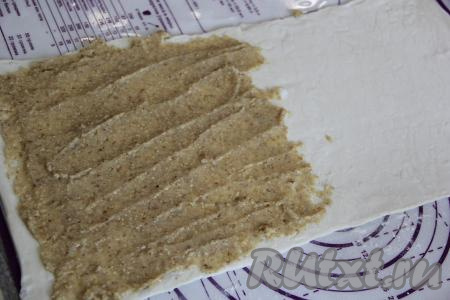 Пласт слоёного теста на припылённой мукой поверхности раскатать в длину до толщины 2-3 миллиметра. Половину раскатанного пласта слоёного теста равномерно смазать ореховой начинкой.