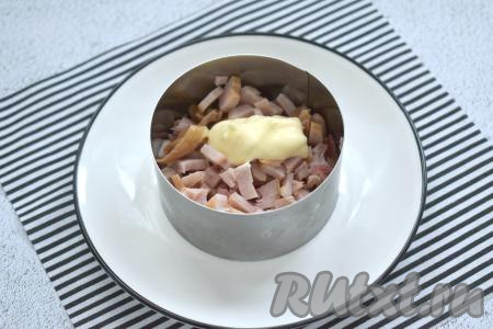 Копчёное куриное мясо нарезаем на маленькие кубики. Ставим каждое сервировочное кольцо на плоскую тарелку. Нарезанное мясо курицы равномерно распределяем по двум сервировочным кольцам, смазываем майонезом, немного утрамбовывая.