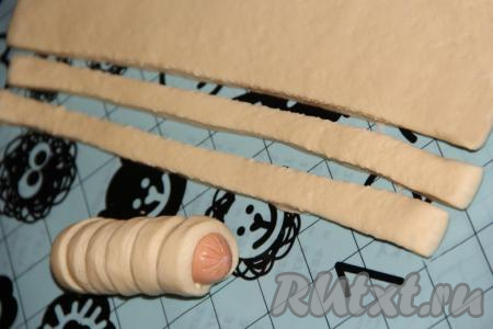 Слоёное тесто для этого рецепта можно взять как дрожжевое, так и бездрожжевое. Тесто нужно заранее разморозить, чтобы оно стало достаточно мягким. Пласты теста выложить на стол (или на силиконовый коврик), слегка присыпанный мукой. Нарезать тесто, не раскатывая, на полоски шириной примерно 0,5-0,8 сантиметра. С сосисок снять плёнку. Обернуть каждую сосиску в полоску из теста. Во время обматывания нужно немного растягивать полоску из теста руками, чтобы она стала более тонкой. Обёртывать сосиску нужно, накладывая полоску из теста немного&nbsp;внахлёст. Хорошо скрепить края полоски. Можно кончики сосисок оставить свободными (без теста). Количество полосок из теста, которое потребуется для обёртывания сосиски, зависит от длины сосисок, которые вы используете.