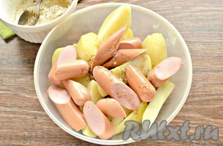 Картофельные дольки и кусочки сосисок выложить в объёмную миску, влить подготовленную смесь масла, чеснока и специй, тщательно перемешать, покрывая картошку с сосисками со всех сторон.