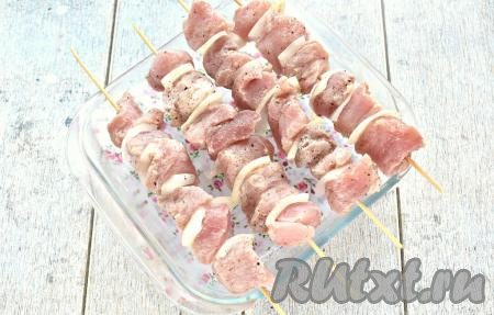 Замаринованные кусочки свинины, чередуя с луком, нанизываем на предварительно замоченные в воде деревянные шпажки. Шашлычки выкладываем на форму с бортиками, как на фото, отправляем в разогретую духовку.