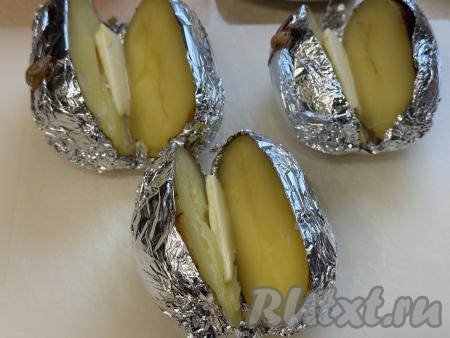 Готовый запечённый картофель переложить на доску. Не снимая фольгу, разрезать каждую картошину на 2 части в длину, не прорезая до конца. Вложить внутрь каждой горячей картошины кусочек сливочного масла. 