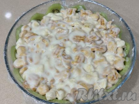 Получившуюся массу из печенья, крема и сметаны выложить в миску с киви, слегка утрамбовать, разровнять, накрыть краями плёнки, свисающими с краёв миски. 