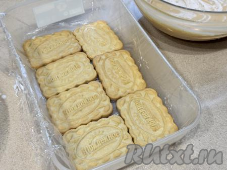 В форму (или в лоток), застеленную пищевой плёнкой, выложить в один слой печенье, пропитанное молоком. Я взяла форму, в которой в один слой помещается, как на фото, 7 штук печенья "Юбилейное". Вы можете взять любую форму, примерив по размеру печенья.