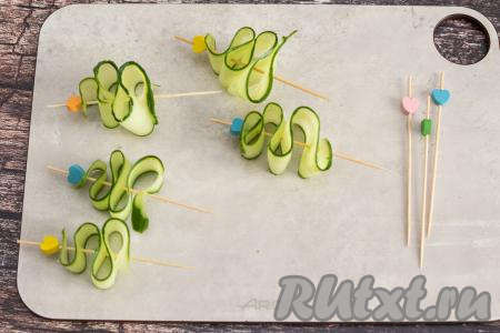 На шпажки нанизать полоску из огурца в виде треугольной спирали, напоминающей внешне ёлочку. Если у вас нет шпажек для канапе, тогда можно взять обычные деревянные зубочистки, а на острый верх каждой зубочистки можно насадить кусочек твёрдого сыра или оливку без косточки.