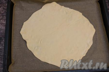 Размороженный пласт слоёного теста на припылённом мукой столе сначала растянуть немного руками, а затем раскатать скалкой до толщины 2 миллиметра. Раскатанное тесто по форме должно напоминать круг. Выложить раскатанный пласт теста на противень, застеленный пергаментом.
