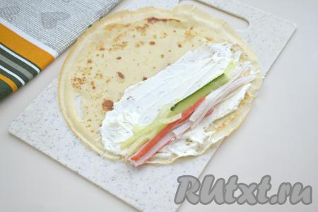 Крабовые палочки вынимаем из упаковки. Крабовые палочки и свежий огурец нарезаем на полоски. Делим зрительно нарезанные палочки и огурцы на 6 равных частей (по количеству блинов). Ближе к одному краю смазанной половины блины выкладываем одну часть крабовых полосок, рядом выкладываем часть огуречных полосок, как на фото.