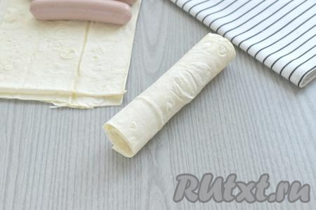 Начиная со стороны с начинкой, плотно заворачиваем сосиску с сыром в лаваш. Конец прямоугольника из лаваша лучше смочить водой и прижать, тогда он не будет загибаться. Аналогично формируем оставшиеся 3 сосиски с сыром в лаваше.