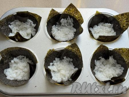 Листы нори нарезать на 6 квадратиков чуть большего размера, чем формы для выпечки маффинов. Каждым квадратиком из нори застелить формочки для маффинов так, чтобы уголки нори торчали из форм. В каждую формочку с нори выложить по 1 столовой ложке отваренного риса, слегка утрамбовать.
