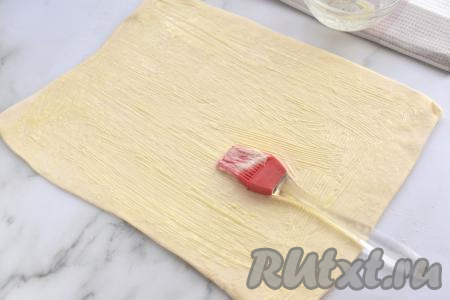 Сливочное масло растапливаем (я растопила сливочное масло в микроволновке в течение 15 секунд), даём ему остыть. Стол присыпаем мукой, выкладываем на него размороженный пласт слоёного теста и раскатываем его в прямоугольник размером ориентировочно 30 сантиметров на 22 сантиметров. Раскатанное тесто с помощью силиконовой кисточки смазываем растопленным сливочным маслом.
