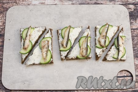 Отрыть банку со шпротами, достать рыбинки. На бутерброды из чёрного хлеба поверх огурца выложить по 1 рыбке.