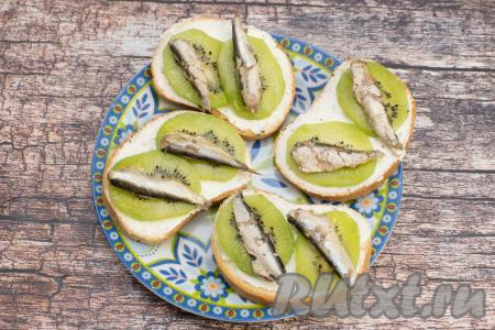 Банку консервированных шпрот открыть и аккуратно достать шпротинки, чтобы они остались целыми. Разложить поверх киви по 2 рыбки на каждый бутерброд.