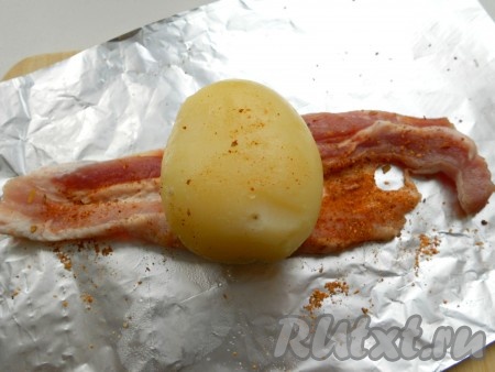 На лист фольги поместить ломтик бекона, посыпать приправой для свинины и паприкой. Сверху выложить целую картофелину и посолить по вкусу.